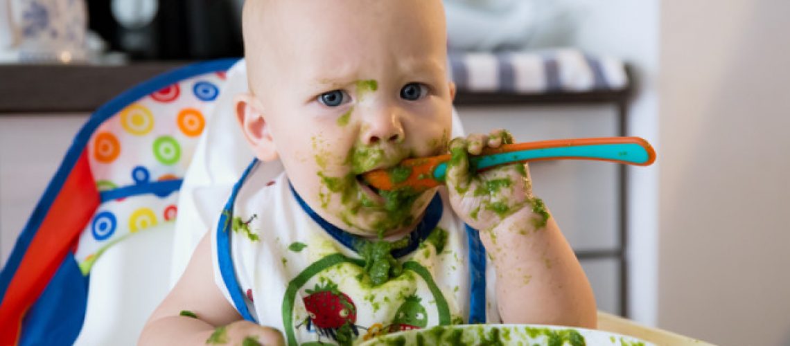 Feeding. Adorable baby child eating with a spoon in high chair. Baby's first solid food